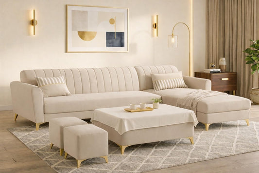 A modern beige sectional sofa with a chaise lounge and matching ottomans in a bright, contemporary living room. The sofa features vertical channel tufting on the backrest and is supported by slim gold-toned legs, set on a patterned rug with a matching coffee table and throw blanket.